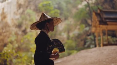 Kimono giymiş, bambu şapkalı, elinde yelpaze tutan bir kadının orta boy fotoğrafı peyzaja bakar ve Kuha Karuhas köşkünün yanında yürür.