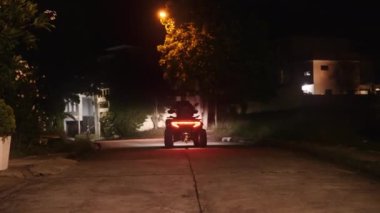 Ağaçlar, evler, sokak lambaları ve arka planda kaldırımları olan yolda ATV kullanan bir adamın videosu.