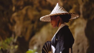 Kimono ve bambu şapkalı bir kadının el vantilatörünü tutarken, bir mağaraya bakarken yan görüntüsü.