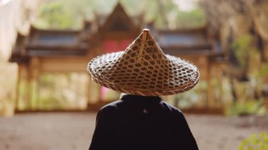Kimono giyen bir kadın, bulanık altın, mavi ve yeşil Kuha Karuhas köşküne bakarken bambu şapkasına dokunur.