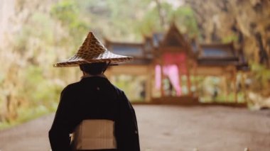 Kimono ve bambu şapkalı bir kadının arka görüntüsü bulanık altın, mavi ve yeşil Kuha Karuhas köşküne doğru yavaşça yürüyor.