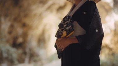 Kimono ve bambu şapkalı bir kadın, bir vantilatör tutuyor, yerinde duruyor ve bir mağaraya bakıyor.