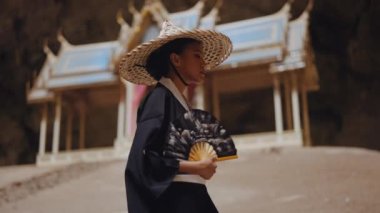 Kimono ve bambu şapkalı bir kadın Kuha Karuha köşkünden çıkarken yelpazesini sallıyor.