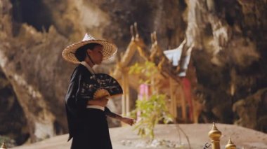 Kimonolu ve bambu şapkalı bir kadın, yelpazesini sallayarak bir mağaranın içinde yürürken Kuha Karuha köşküne bakıyor.