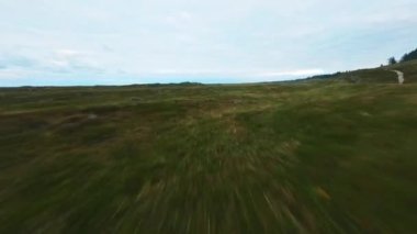 Bulutlu bir günde, deniz kenarındaki çimenli bir tepe üzerinde alçaktan ve hızlı uçan bir dronun görüntüsü.