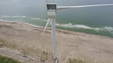 Bulutlu gökyüzünün altında, kumsalda rüzgar türbininin üzerinden uçan bir dronun gündüz FPV görüntüsü.