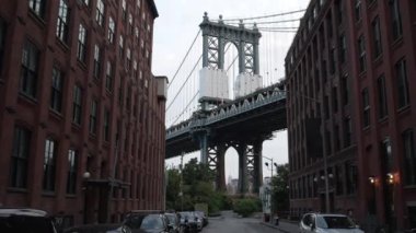 Eğik bir çekim aşağı doğru hareket ediyor. Manhattan köprüsünün, DUMBO mahallesindeki iki bina ile gündüz vakti tuğlalı kaldırımda duran sabit arabalar arasında çerçevelendiğini gösteriyor.