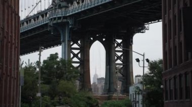 Manhattan Köprüsü 'nün yukarı doğru hareket ettiğini gösteren eğik bir çekim. Gündüz saatlerinde DUMBO mahallesindeki iki bina ve durgun arabalar arasında çerçevelenmiş.