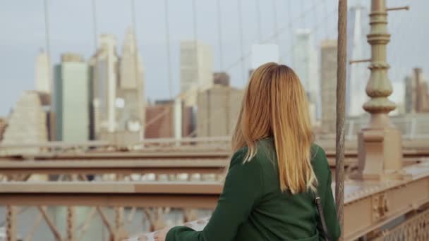 昼間にマンハッタン橋とニューヨークのスカイラインを望むブルックリン橋で緑のコートを着た女性の背中の景色を示すビデオ — ストック動画