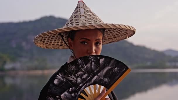 着物姿の女性の中近接撮影では カメラに焦点を当て 顔の近くに手のファンを置き タイの日没時に竹の帽子に触れながら景色を眺めます — ストック動画