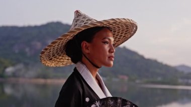 Kimono giyen, bambu şapkalı, el fanı tutan bir kadının orta boy yakın çekimi kameraya odaklanıyor ve Tayland 'da gün batımında arka planda bulanık dağ manzaralı göl tapınağına uzaktan bakıyor.