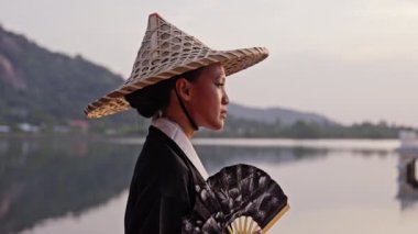 Kimono giyen, bambu şapkalı, el fanı tutan bir kadının orta ölçekli yakın çekimi. Tayland 'da gün batımında arka planda göl tapınağı ve dağ manzarası olan kameraya odaklanıyor ve diğer tarafa bakıyor.