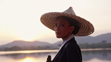 Kimono ve bambu şapka takan, kameraya bakıp gülümseyen, yüzünün yanındaki pervaneyle uzaklaşan ve gün batımında Tayland 'dan ayrılan bir kadının orta boy yakın çekimi.