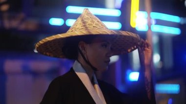 Kimono giymiş, aşağı bakan bir kadının orta boy yakın çekimi, bambu şapkasına dokunur ve yukarı bakmaya başlar, gece boyunca mavi ve sarı neon ışıklı bulanık arka plana doğru.