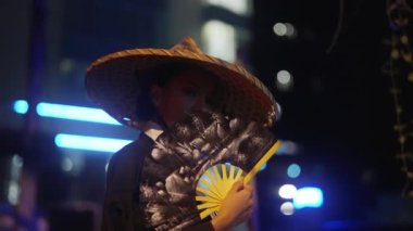 Kimono ve bambu şapkalı bir kadın kameraya bakıyor ve ağzını el vantilatörüyle örtüyor, sonra da bulanık arkaplana, mavi ve beyaz neon ışıklarla doğru bakmaya başlıyor.