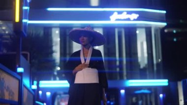 Kimono ve bambu şapkalı bir kadın, bir el yelpazesi tutuyor, sağ tarafından yürüyor ve bakıyor, sonra gece vakti mavi neon ışıklarıyla bulanık arka plana doğru dönüyor.