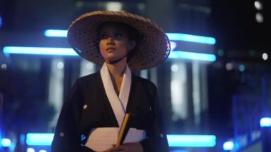 Kimono ve bambu şapkalı bir kadın, sağ tarafına bakarken yürür sonra da gece vakti mavi neon ışıklarıyla bulanık arka plana doğru ilerler.