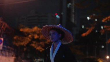 Kimono ve bambu şapkalı bir kadının orta ölçekli bir yakın çekimi. Önüne bakıp, ağaçlar ve binalarla kaplı bulanık bir arka planda, sıcak beyaz bir sokak lambasının önünden geçerken yürüyor.
