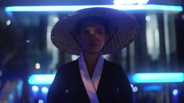 Kimono ve bambu şapkalı bir kadın, yüzünde sıcak beyaz ışık ve ağaç gölgeleriyle yürüyor, etrafına bakıyor, sonra da bulanık binaya karşı mavi ışıklı bir dönüş yapıyor.