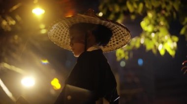 Kimono ve bambu şapkalı bir kadın, pervanesine bakıyor, sonra kameraya, bulanık beyaz sokak ışıkları, stop lambası, ve arka plandaki ağaçlar gece boyunca