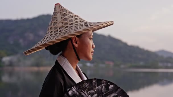 在泰国 一名身穿和服 头戴竹帽 手持扇子的女子在日落时望着朦胧的高山和湖景 拍了一张中等特写 — 图库视频影像