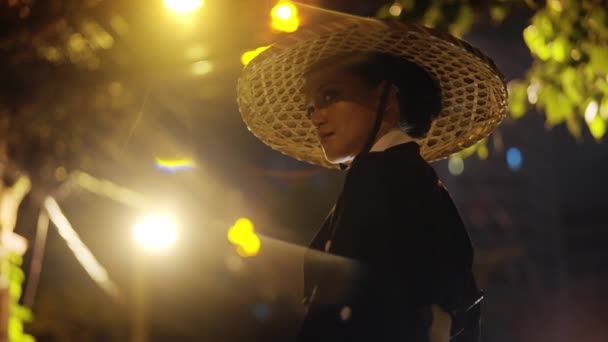 身穿和服 头戴竹帽的女人 左顾右盼 把扇子放在左肩上 并在夜色朦胧的温暖的街灯下向右张望 — 图库视频影像