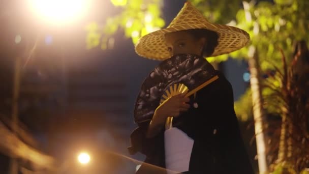 着物と竹の帽子を被った女性は カメラを見て 暖かい白い街路灯 夜の植物を背景に 手のファンで彼女の口を微妙に覆います — ストック動画