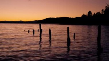 A video of the calm waters of the lake in Bariloche, Argentina showing the silhouette of the mountain from afar, and logs floating vertically in place on the center of the frame, during sunrise