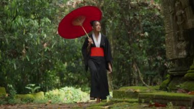 A full shot of a woman wearing a kimono, walking on the mossy temple grounds in Cambodia during daytime