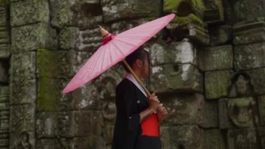 A medium shot showing the side view of a woman wearing a kimono, walking near the mossy temple stone walls in Cambodia during daytime