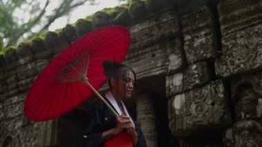 A medium shot of a woman wearing a kimono, gazing while walking at the mossy temple ruins in Cambodia during daytime