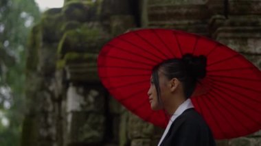 A medium close-up shot showing the side view of a woman wearing a kimono, looking down while walking at the mossy temple ruins in Cambodia during daytime
