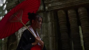 A medium shot of a woman wearing a kimono, visiting the mossy temple ruins during daytime in Cambodia