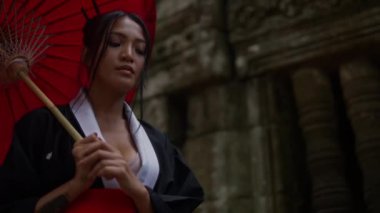 A medium close-up shot of a woman wearing a kimono, walking around the mossy temple ruins during daytime in Cambodia
