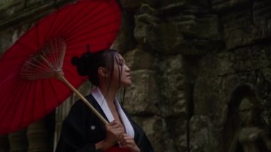 A medium close-up shot of a woman wearing a kimono, walking and looking at the walls of the mossy temple ruins during daytime in Cambodia
