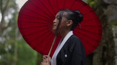 A medium shot of a woman wearing a kimono and holding a red wagasa, looks at the camera at Ta Prohm temple located in Cambodia