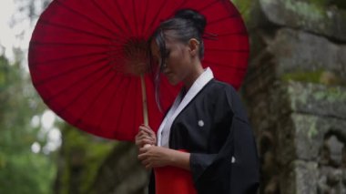 A medium shot of a woman wearing a kimono and holding a red wagasa, looks at the camera at Ta Prohm temple located in Cambodia