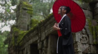 A medium full shot of a woman wearing a kimono and holding a red wagasa at Ta Prohm temple looking towards the camera and faces away at the end