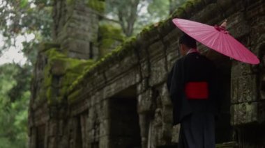 A video showing the back view of a woman wearing a kimono, walking at Ta Prohm temple during daytime in Cambodia