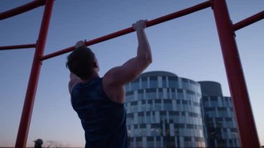 A back view of a man doing a calisthenic exercise called pull-ups on a rooftop during sunset