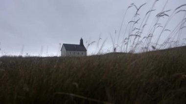 View from the ground of a man standing outside the Strandarkirkja church while the grass sways in the wind
