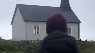 The camera pans from the back of a man wearing a jacket and a beanie, standing outside the Strandarkirkja church during daytime in Iceland