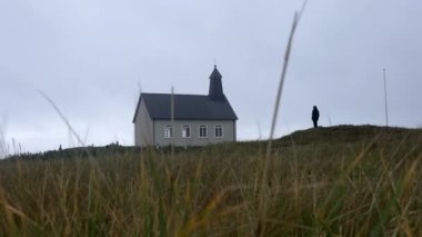 A man stands outside the Strandarkirkja church as the camera gently tilts downwards showing the grass swaying in the wind