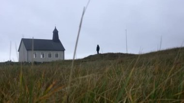 View of a man standing on the ground in front of the Strandarkirkja church as the camera gently moves through the wind-blown grass