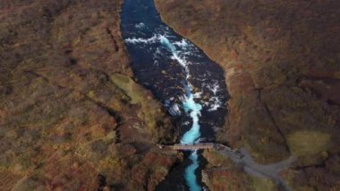 Aerial drone view of the blue water stream of the Bruarfoss waterfall and the wooden bridge above on the riverside's green landscape