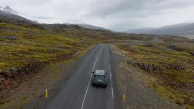 A wide shot of a grey car travelling on a mountain pass road with a view of a snowy mountainside in Iceland during daytime