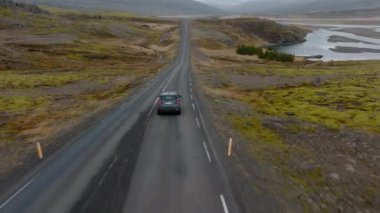 An aerial shot of a grey car venturing on a mountain pass road with a view of a river on the right side and a snowy mountain landscape, in Iceland during daytime