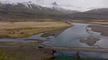 A wide shot of a grey car travelling on a bridge over a river, with a view of a snowy mountainside in Iceland during daytime