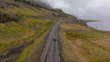 An aerial shot of a grey car venturing on a mountain pass road with a view of a river on the right side, in Iceland during daytime