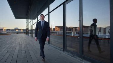 A full shot to medium shot of a businessman walking in a straight direction with his reflection showing on the glass facade during daytime